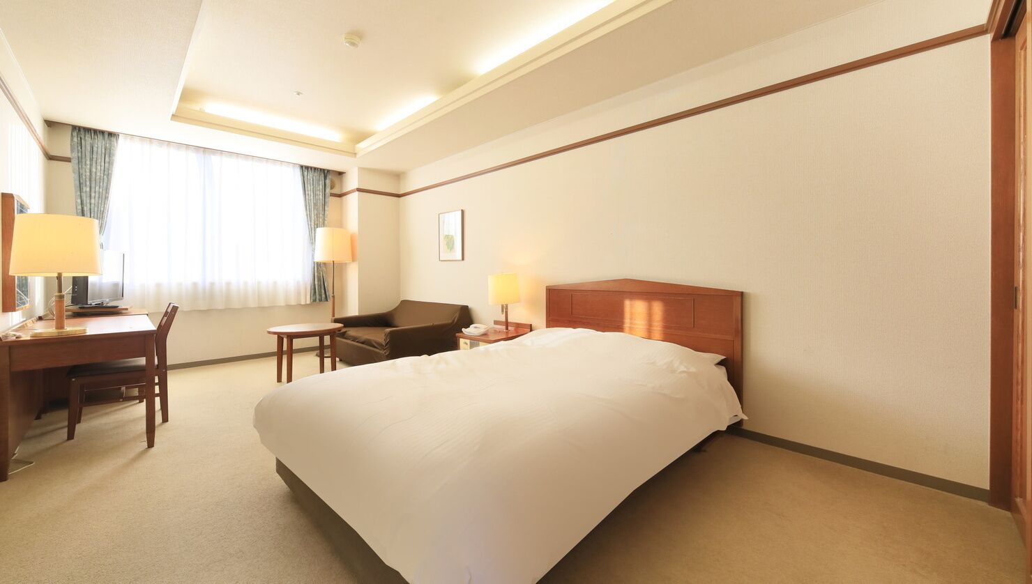A hotel room with a large bed, a desk with a chair, a small table, two lamps, and a window with curtains.