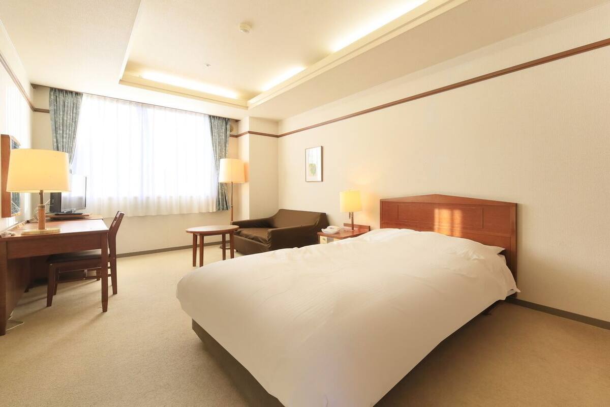 A hotel room with a large bed, a desk with a chair, a small table, two lamps, and a window with curtains.