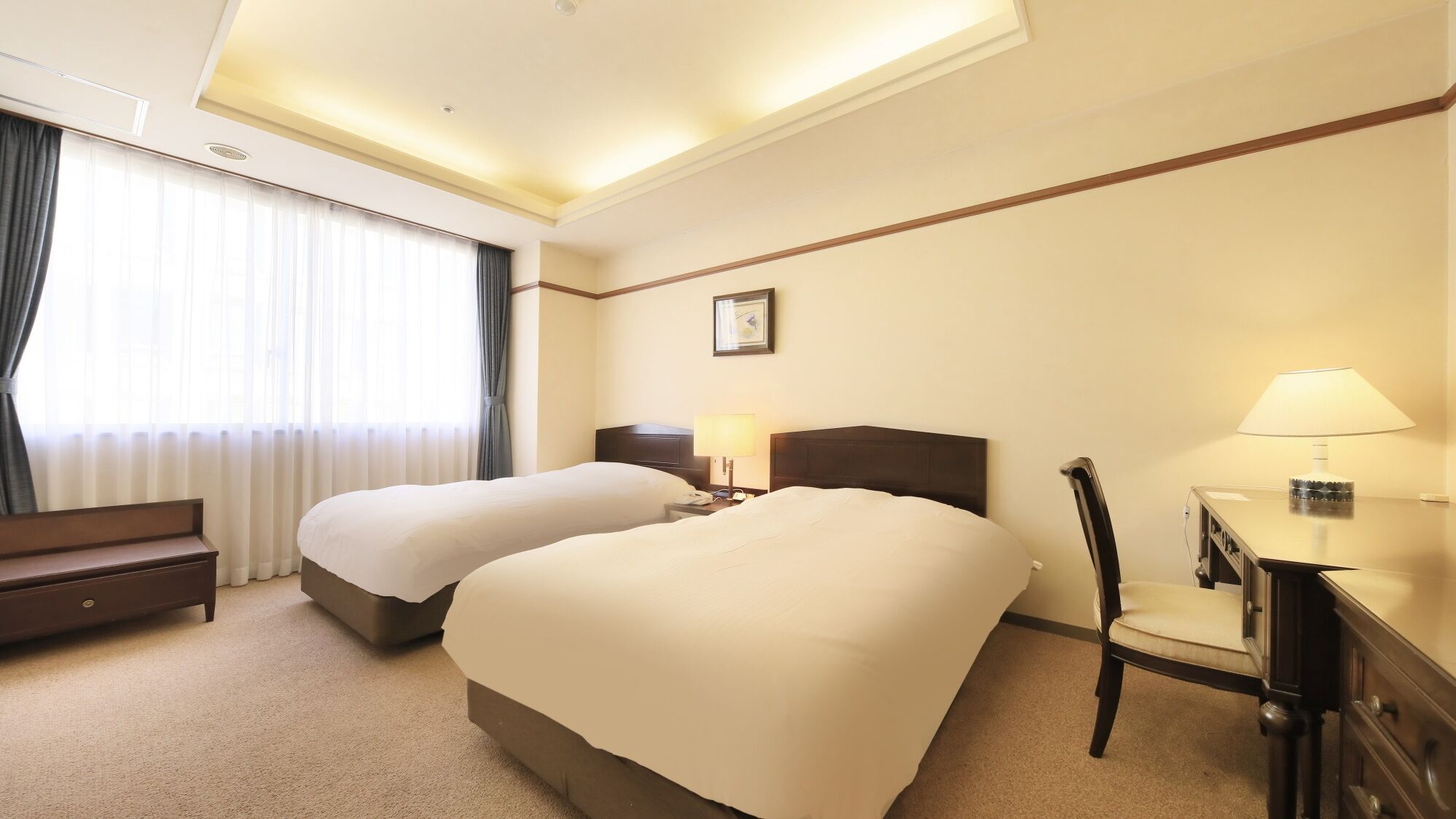 A hotel room with two beds, a desk, a chair, and a nightstand with a lamp.