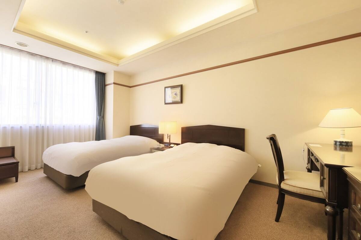 A hotel room with two beds, a desk, a chair, and a nightstand with a lamp.