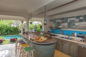 A modern kitchen with a curved island, bar stools, and a view of a swimming pool. - Villa Sky Li By Nagisa Bali (Seminyak)