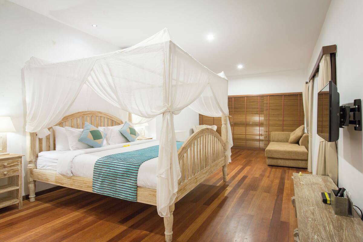 Une chambre avec un lit sous un baldaquin, une tête de lit en bois, une table de chevet, un téléviseur fixé au mur et un canapé dans un coin.