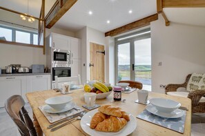 A kitchen with white cabinets, a microwave, and an oven. There's a dining table with plates, a bowl of fruit, and a croissant.