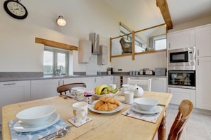 Une cuisine moderne avec des meubles blancs, une table à manger en bois mise avec des assiettes, des bols et un croissant, et une horloge au mur.