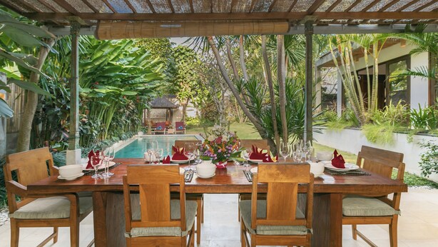 An outdoor dining area with a pool view, wooden furniture, and tropical plants.