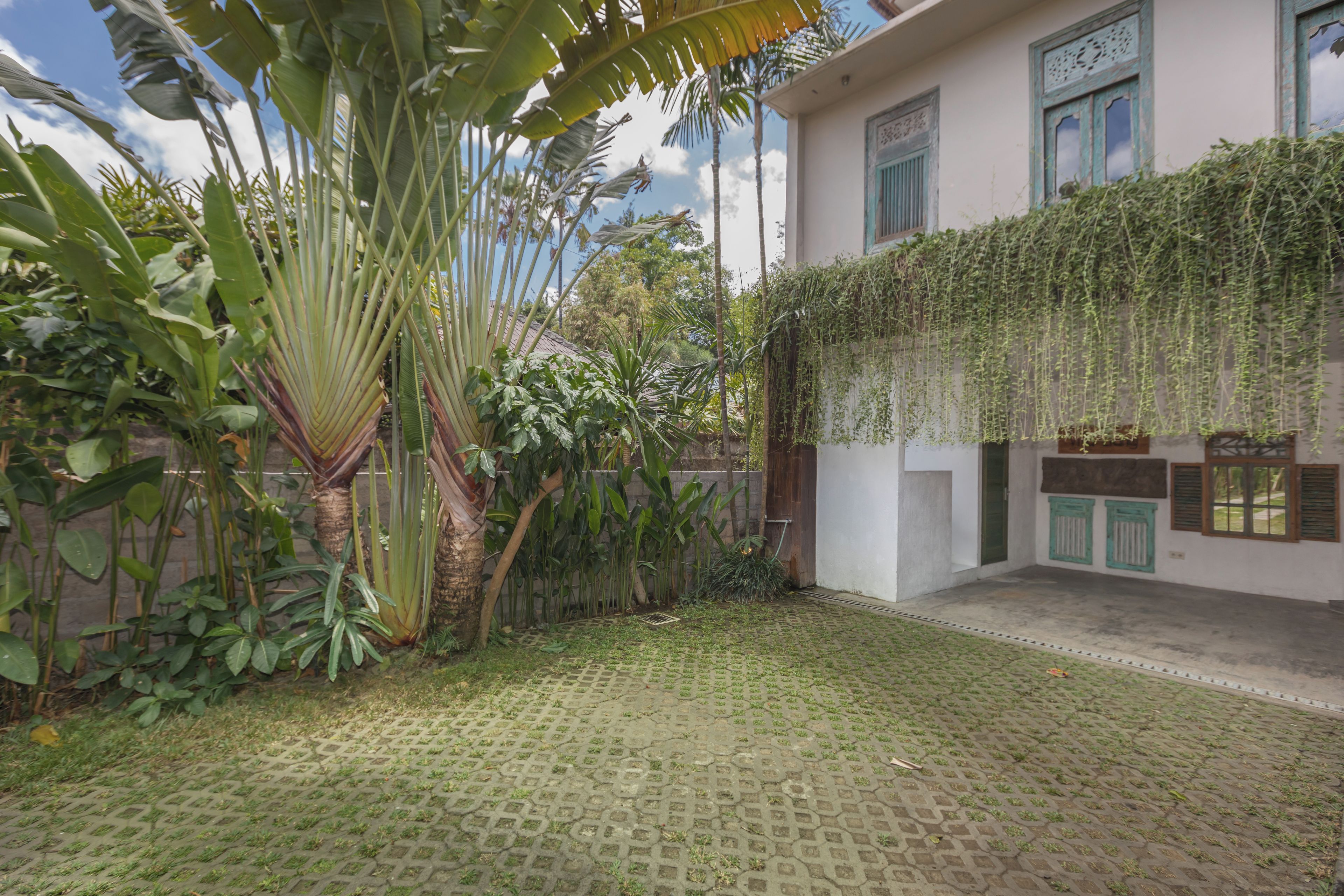 A courtyard with a paved floor, surrounded by lush greenery and a building with a white exterior and green shutters.