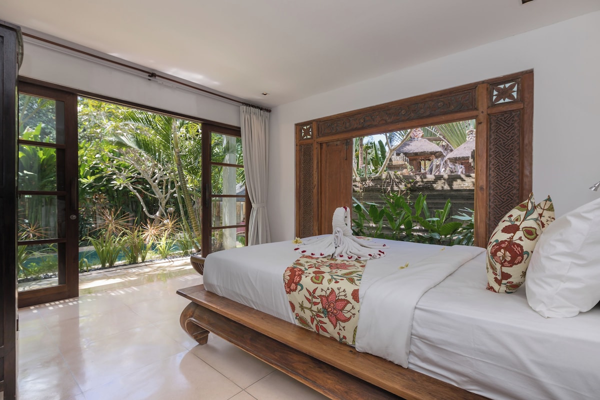 Quarto com uma cama grande, vista para vegetação exuberante e detalhe no teto de madeira.