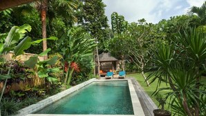 A rectangular pool with a wooden deck in the foreground, surrounded by lush tropical vegetation and a small gazebo with blue chairs in the background.