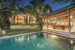 A poolside area with a dining setup and a building with large windows and a covered patio. - Villa Liang by Nagisa Bali (Seminyak)