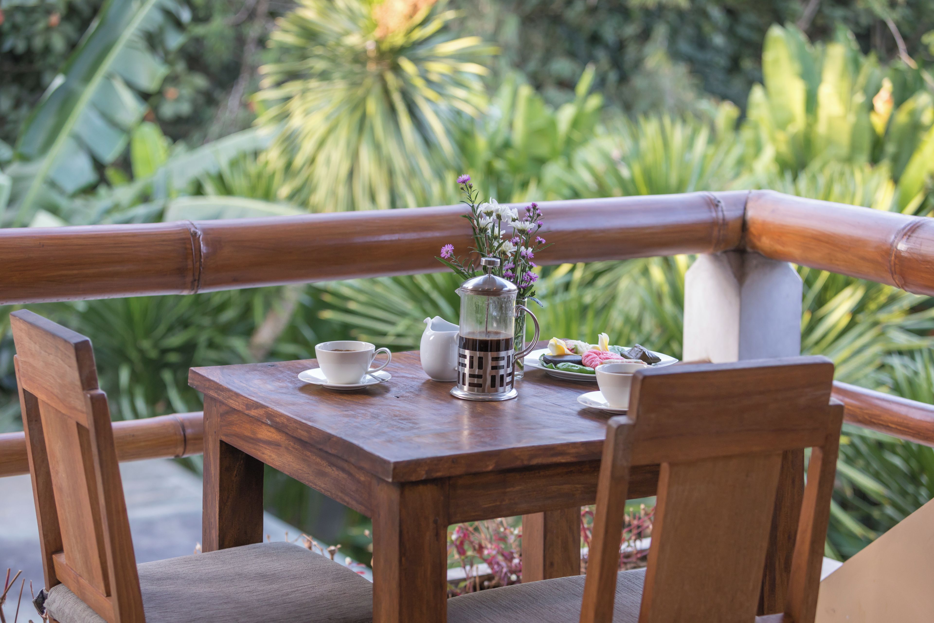 A wooden table with a tea set, a pitcher, and a plate of food, set against a backdrop of lush greenery.