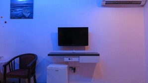 Habitación con televisor de pantalla plana sobre un soporte negro, un mini refrigerador blanco, una silla de madera y aire acondicionado instalado en la pared.