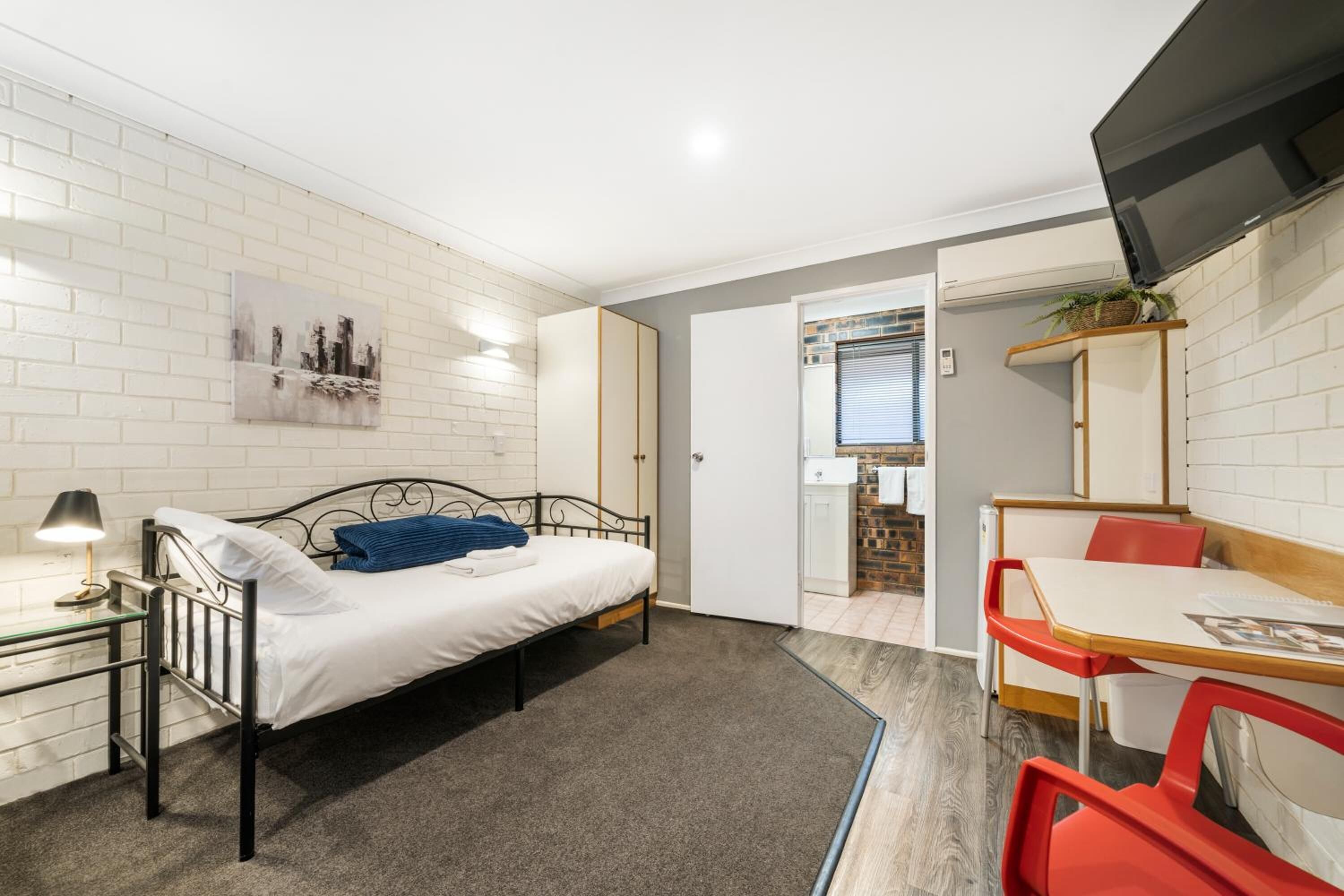 A compact room with a bed, a desk, and a chair. There is a TV mounted on the wall and a door leading to a bathroom.