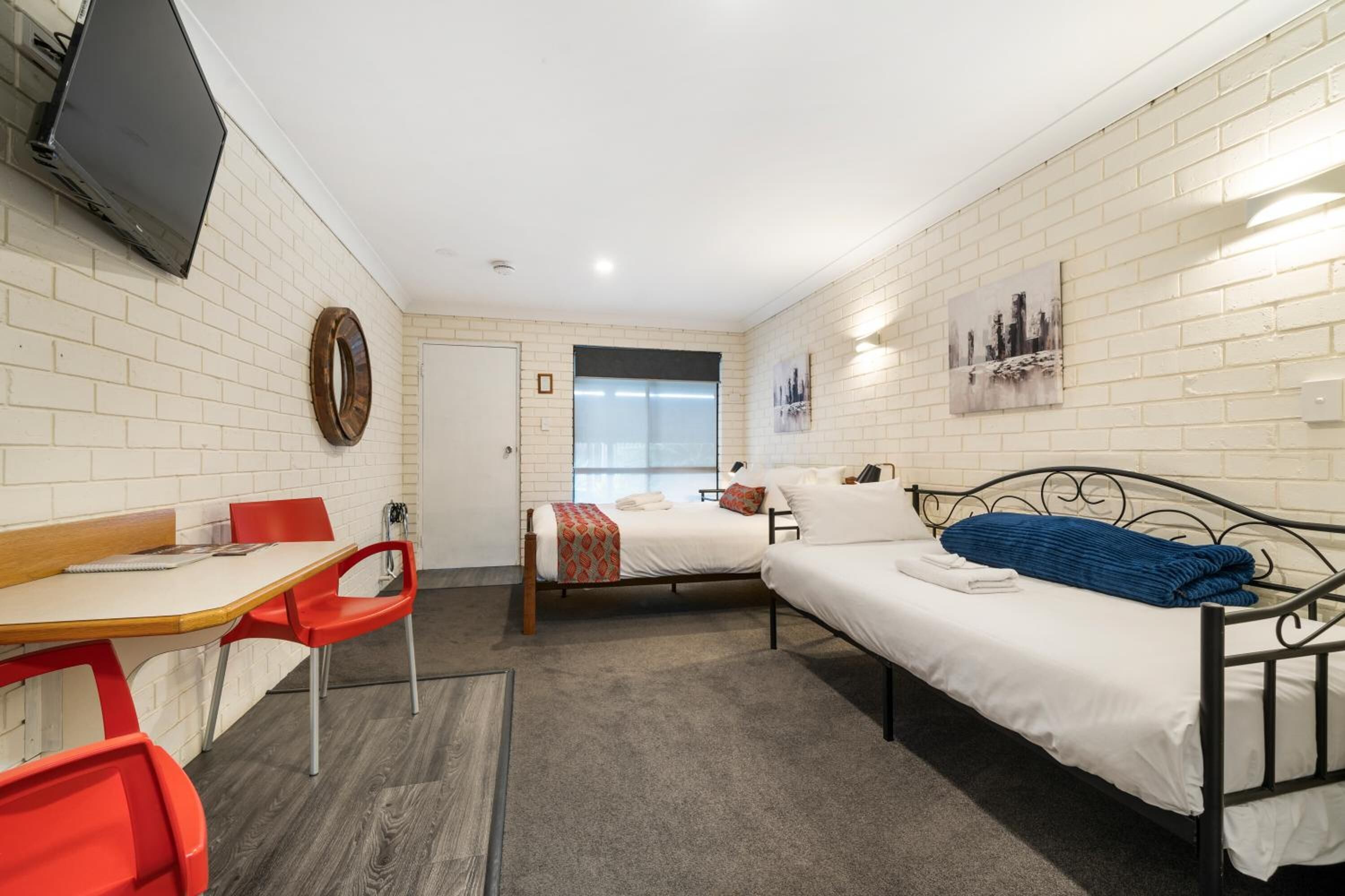 A small, well-lit room with two beds, a desk, and a TV.