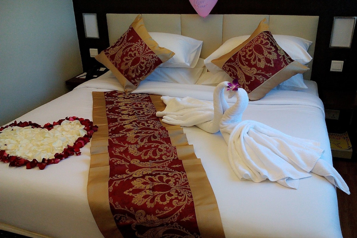 Una cama bien hecha con un arreglo de flores blancas y rojas en forma de corazón, un corredor de cama con estampado rojo y dorado, y dos toallas blancas dispuestas en forma de cisne.
