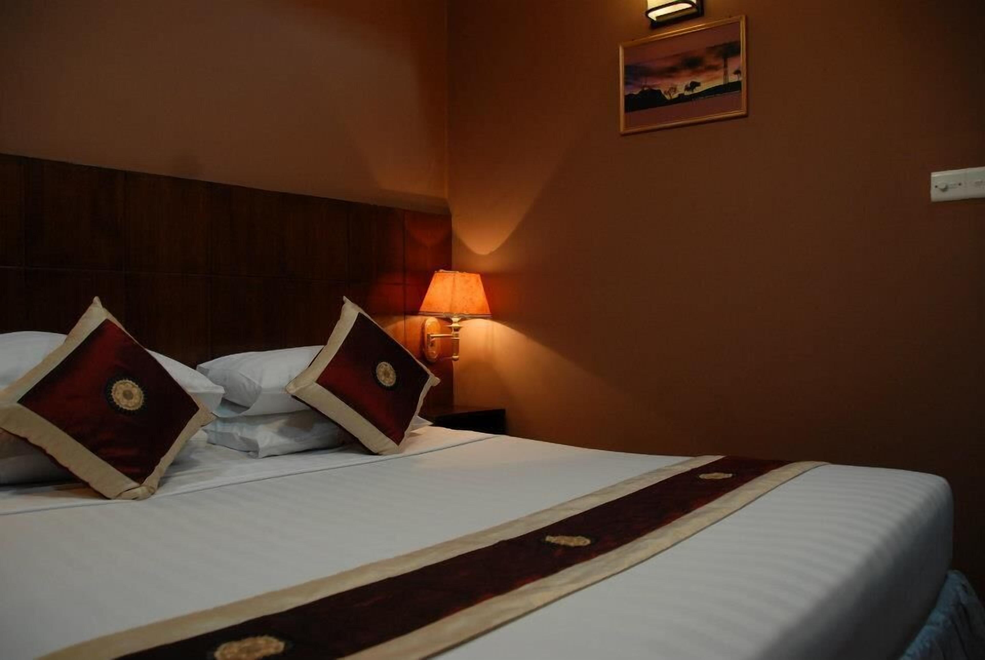 A neatly made bed with white linens and two decorative pillows, a wooden headboard, a bedside lamp, and a framed picture on the wall.