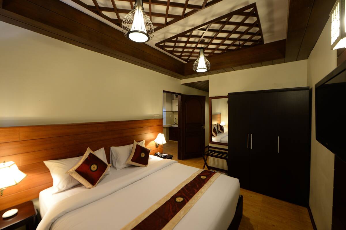 Una habitación de hotel con cabecera de madera, una cama grande con ropa de cama blanca y almohadas decorativas, una mesita de noche con lámpara y un armario.