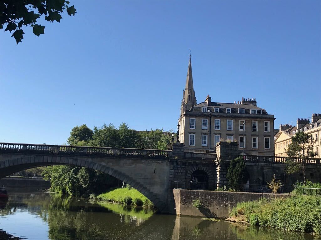 A stone bridge over a calm river, with a large building featuring a tall spire in the background.