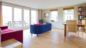 A spacious living room with a blue sofa, a red armchair, a wooden dining table, and a bookshelf. - Charming reed roof Lakehouse, with surrounded garden, trampoline and watersports option (Haren)