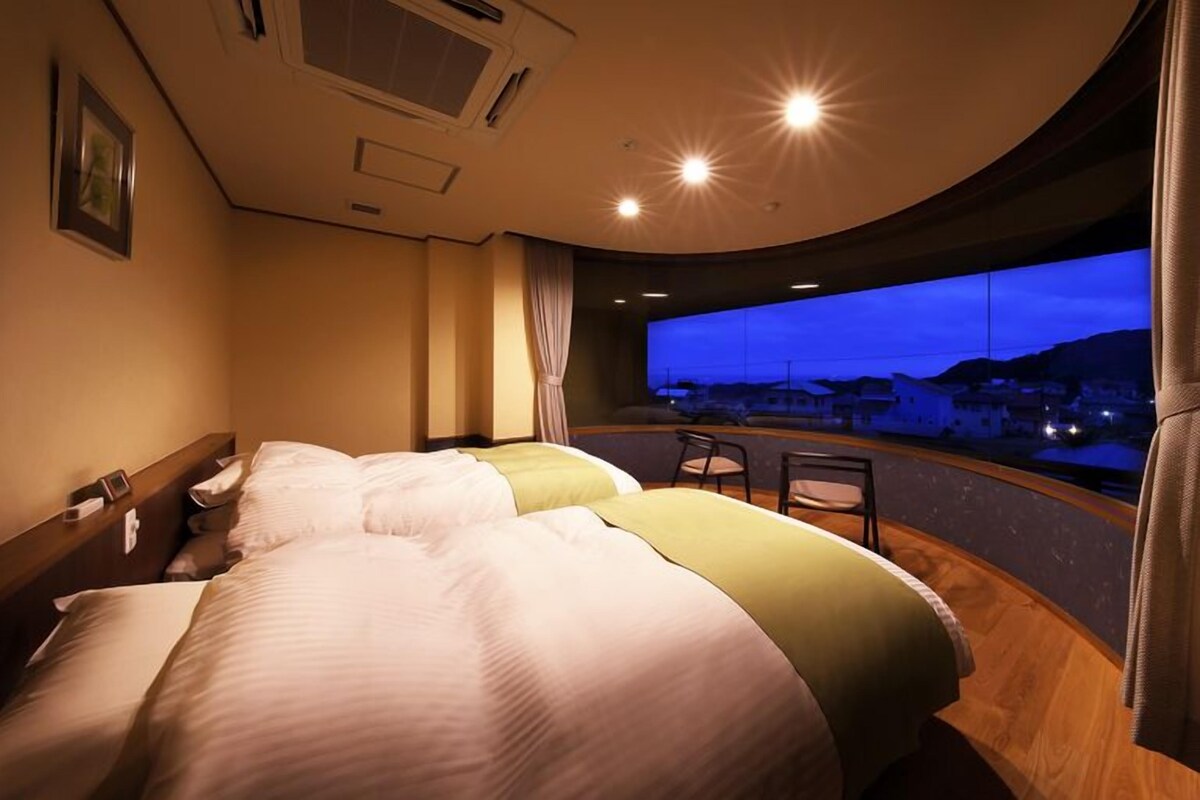 Une chambre d’hôtel avec deux lits, une vue sur la ville au crépuscule et un balcon avec des chaises.