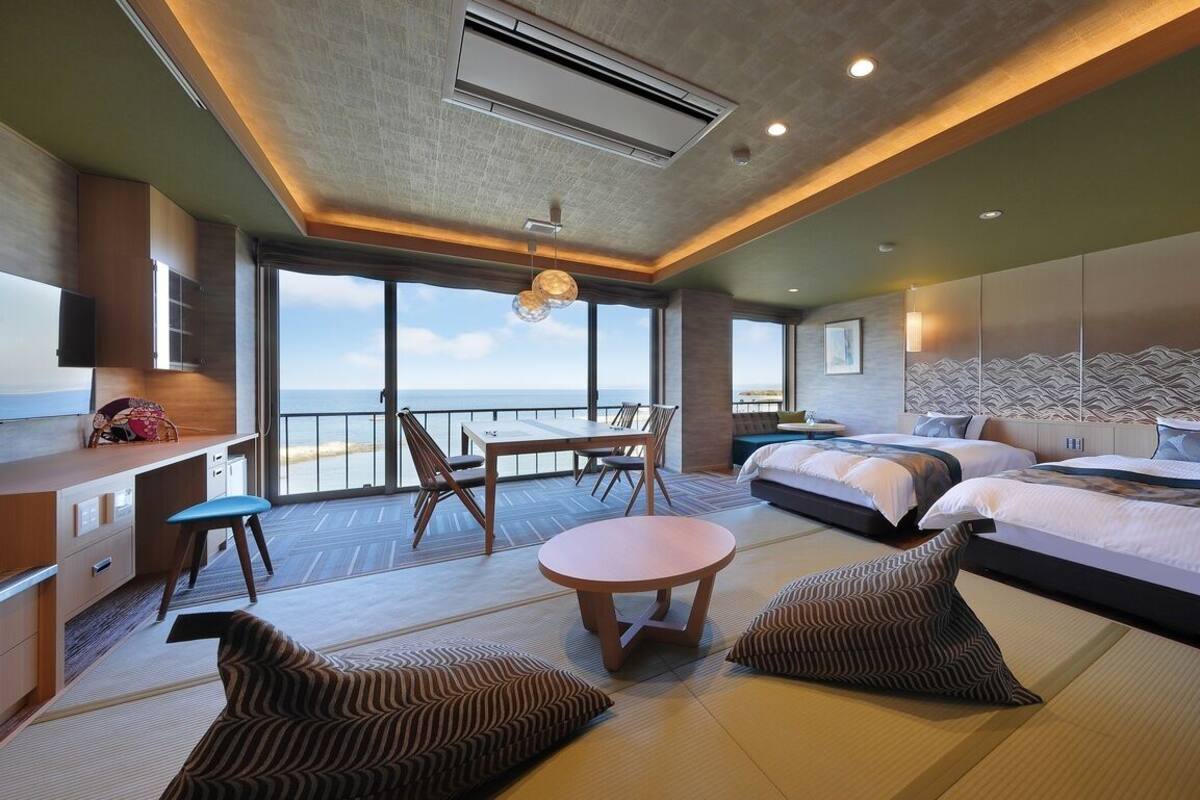 Habitación de hotel con balcón, zona de comedor, dos camas, escritorio y ventana grande.