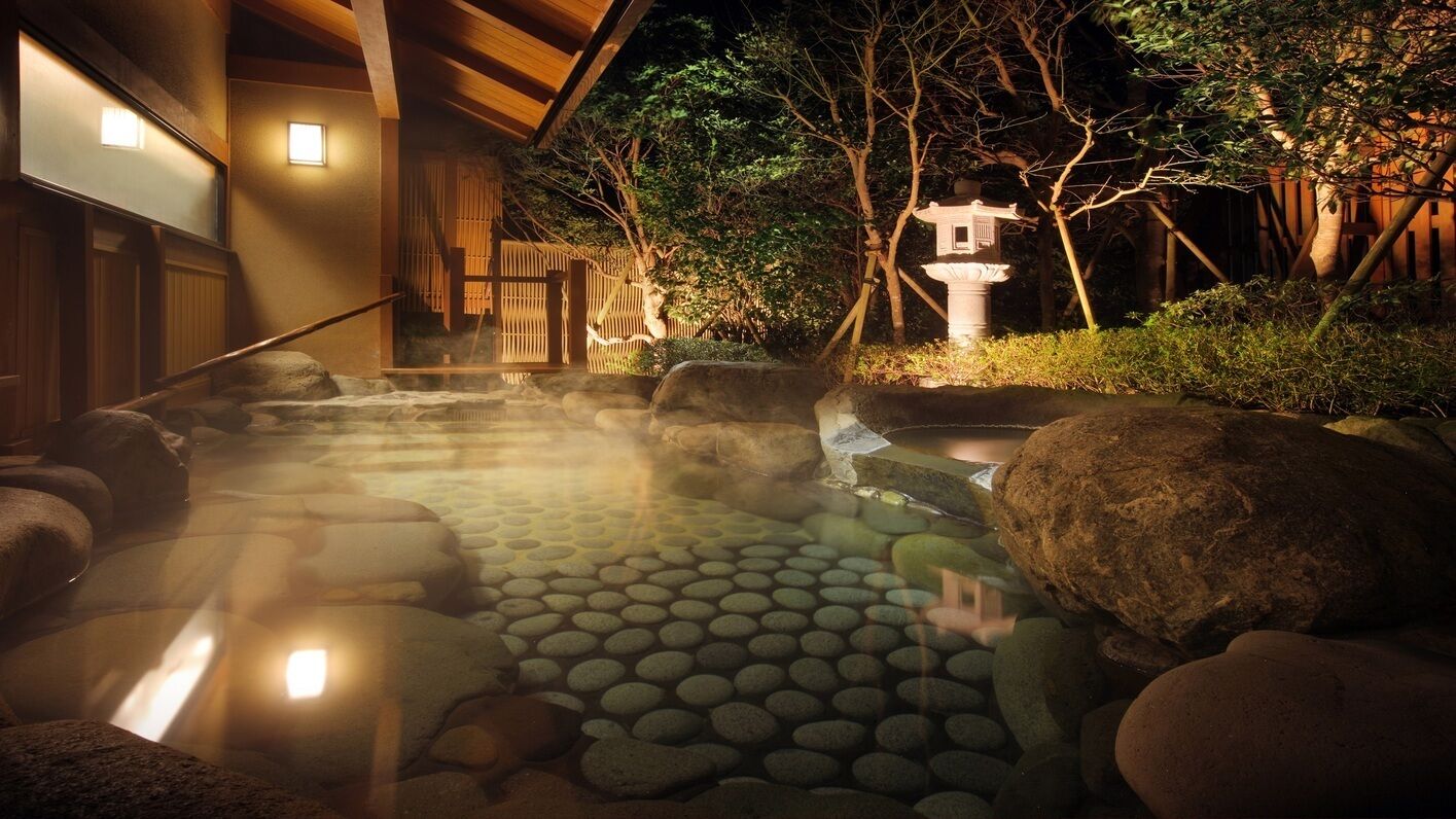 A traditional Japanese onsen with a stone bath surrounded by rocks, a wooden structure, and a small shrine.