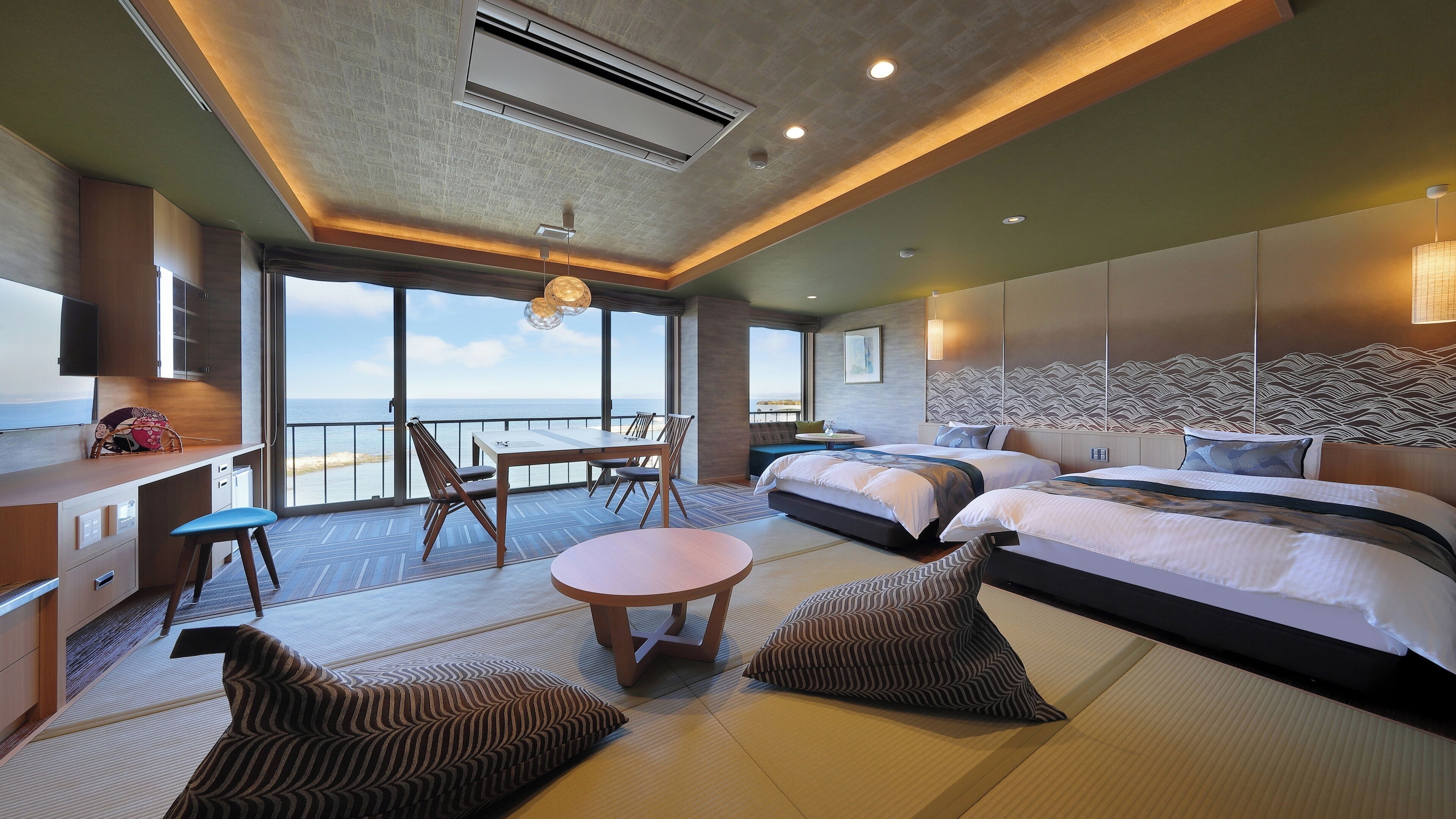 A modern hotel room with a large bed, a desk, and a dining area with a view of the sea.