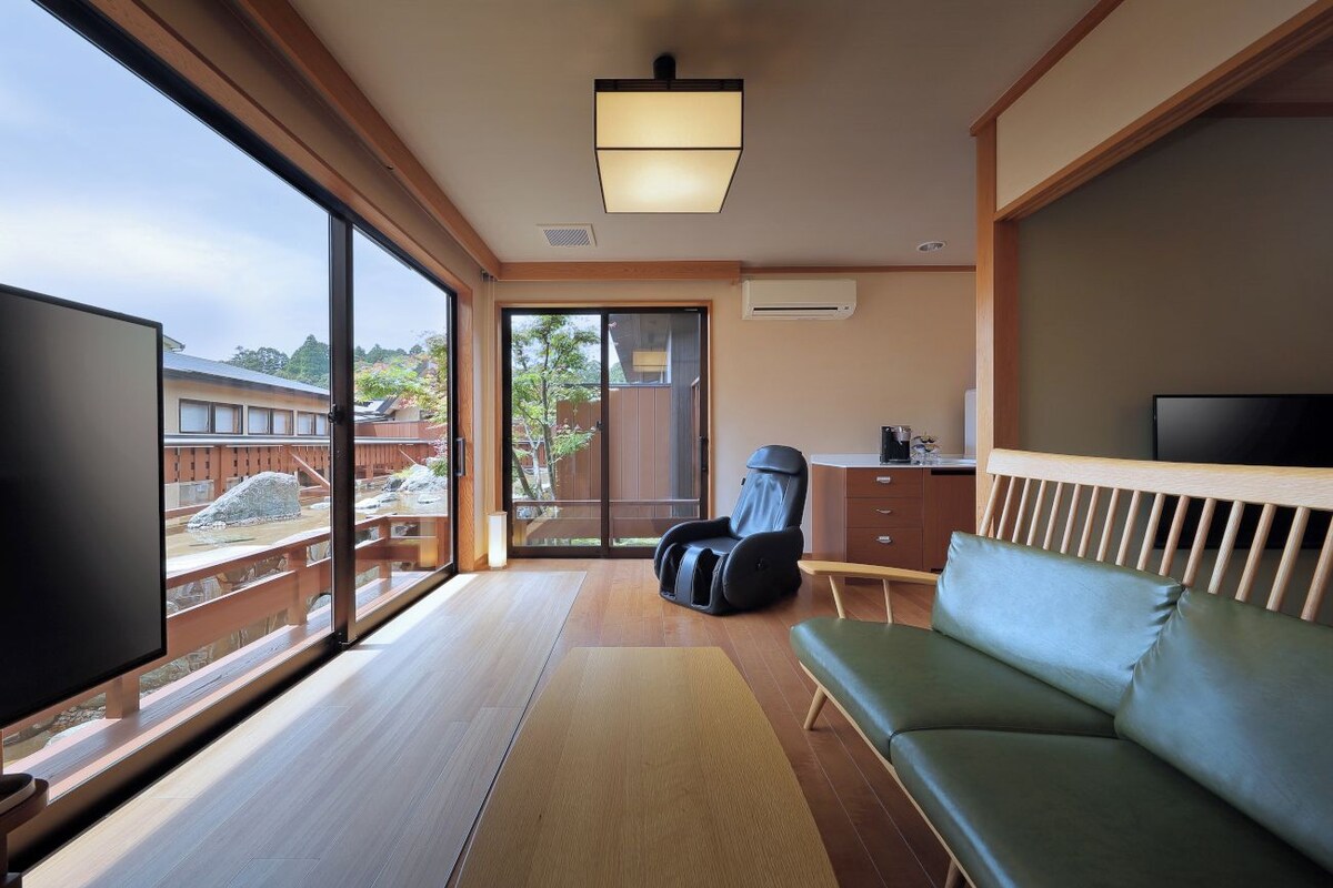 A room with a large window, a massage chair, a sofa, a flat-screen TV, and a wooden ceiling.