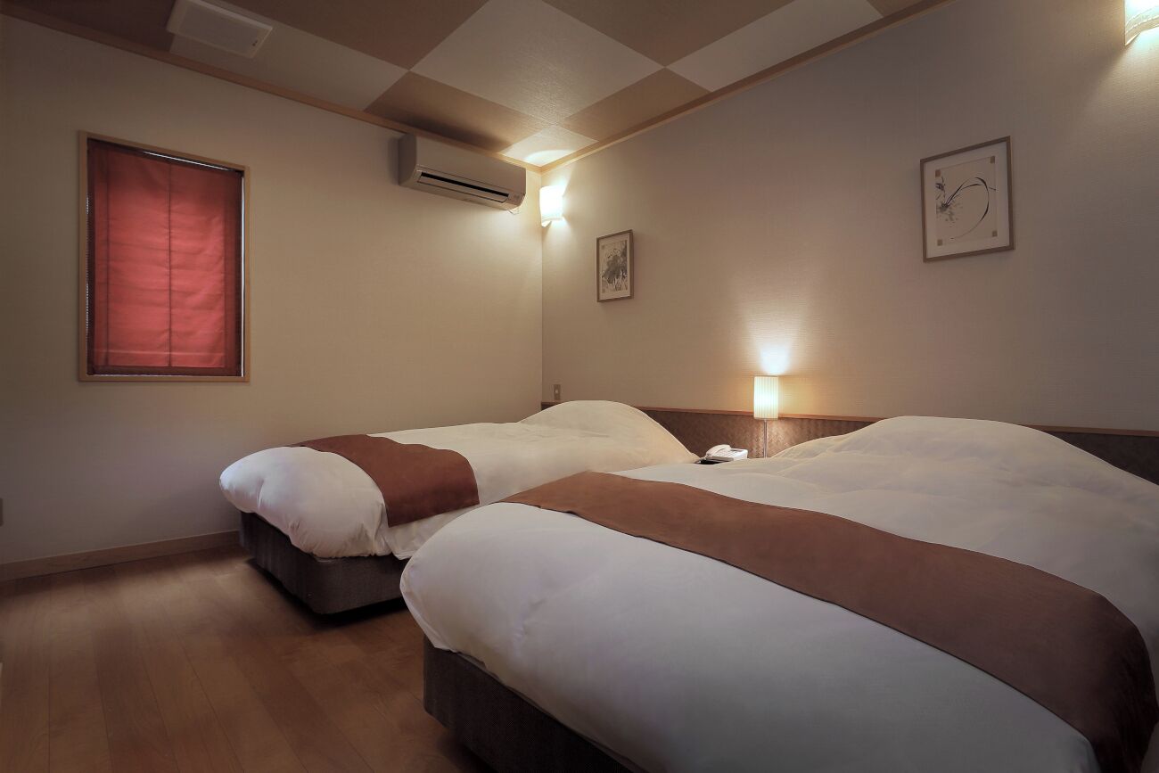 A hotel room with two beds, a window with blinds, an air conditioning unit, a wall-mounted light, and a framed picture.