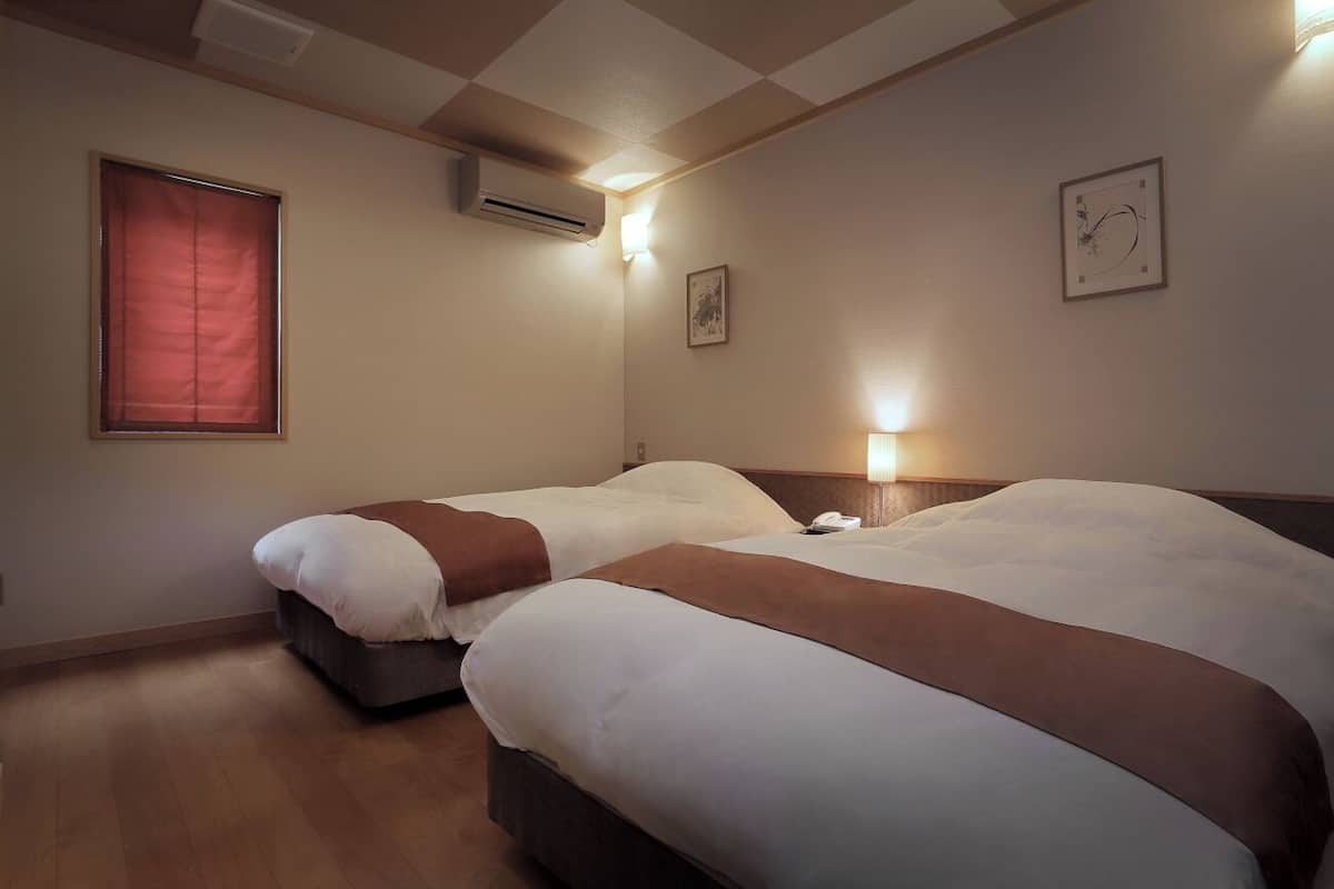 A hotel room with two beds, a window with blinds, an air conditioning unit, a wall-mounted light, and a framed picture.
