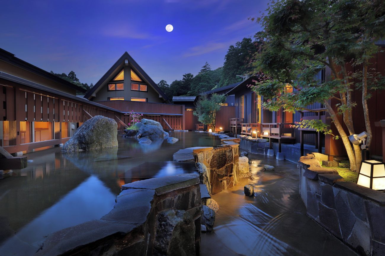 Un ryokan japonés tradicional con un manantial de piedra caliente, edificios de madera y un cielo nocturno con luna visible.
