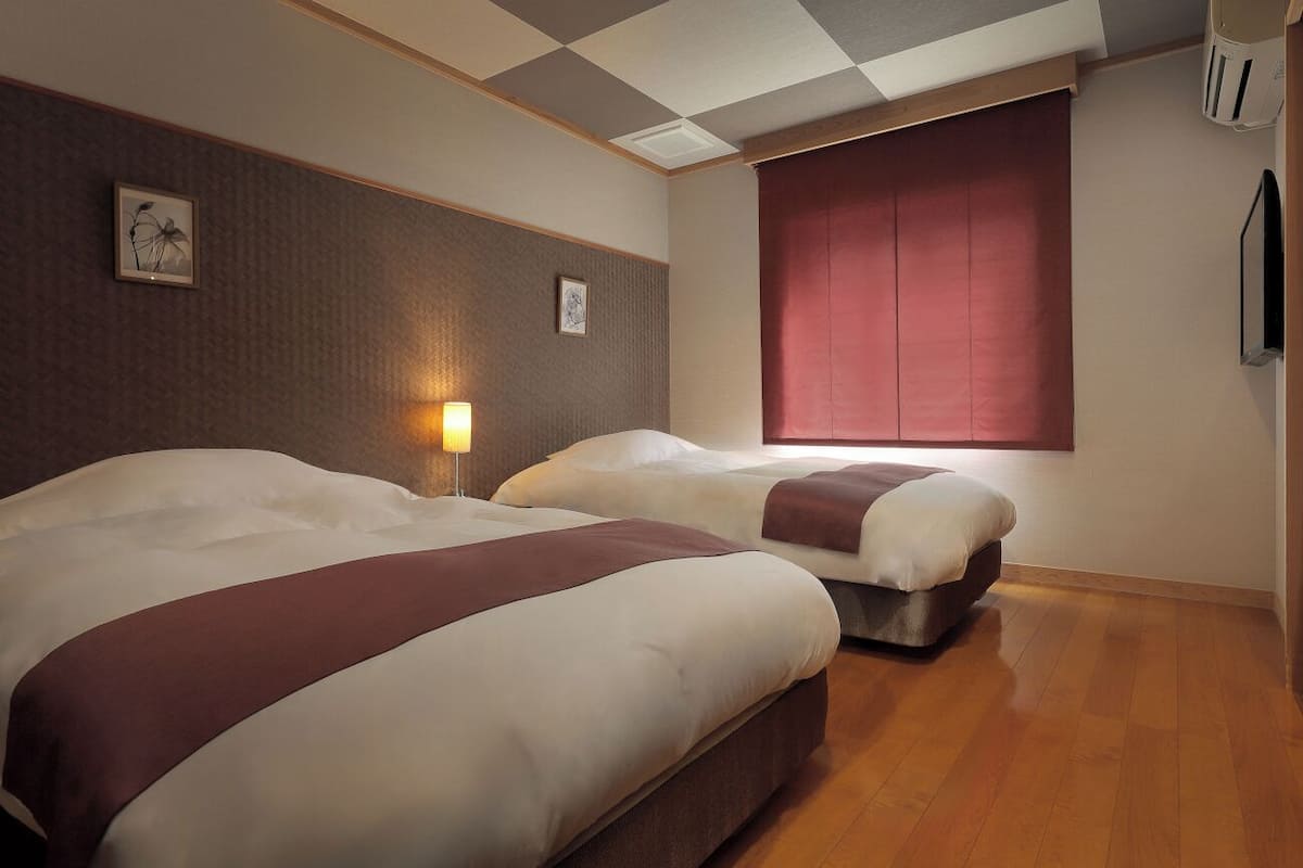 A hotel room with two beds, a window with red curtains, a wall-mounted lamp, and a framed picture.