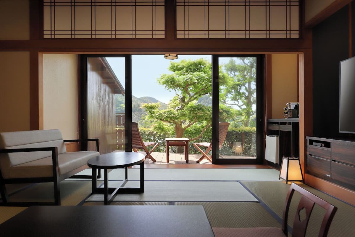 A room with tatami flooring, a sofa, a table, a TV, and a sliding door leading to a balcony with a view of trees and mountains.