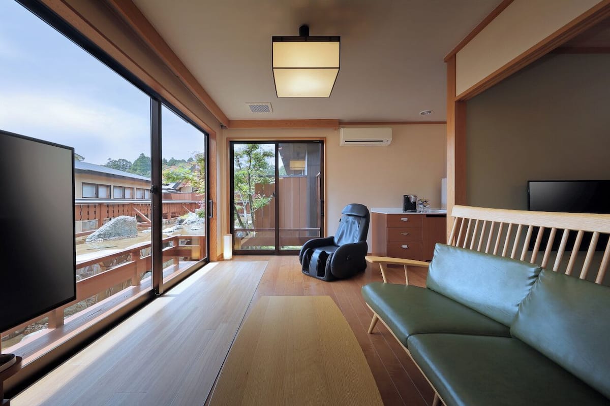 A room with a large window, a massage chair, a sofa, a flat-screen TV, and a wooden ceiling.