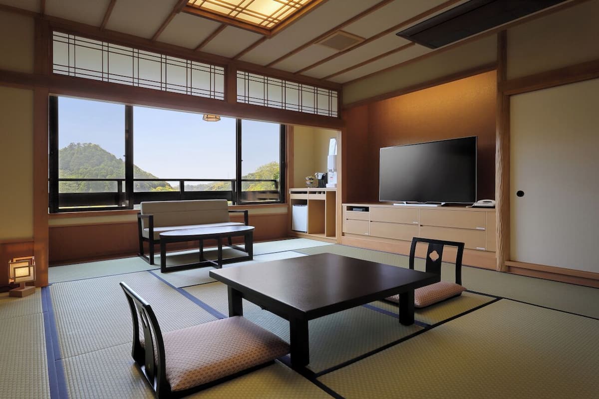 A traditional Japanese room with tatami flooring, a low table, seating, a television, and a view of a mountain.