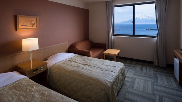 A hotel room with two beds, a desk, a chair, a lamp, a window with a view of mountains and the sea, and a painting on the wall. - Mitsui Kanko Hotel (Rebun)