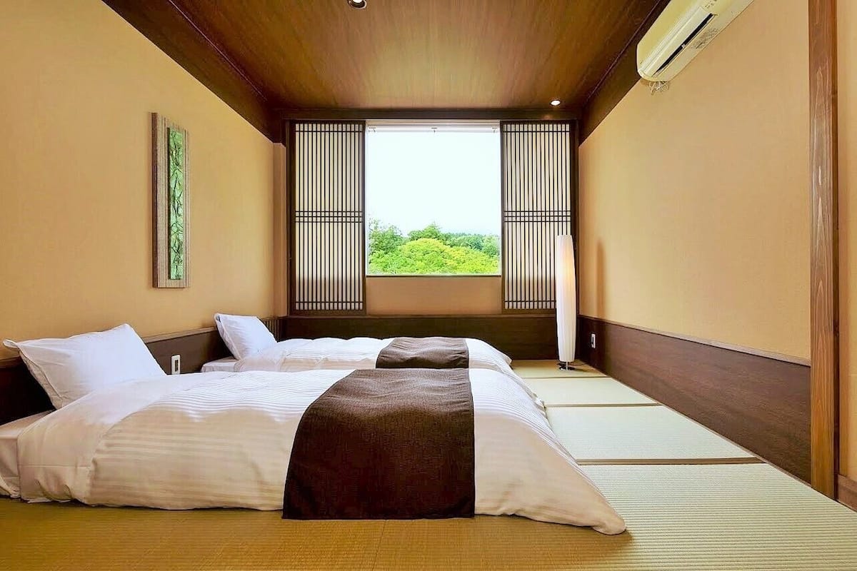 A hotel room with tatami mats, a bed with white and brown bedding, a window with a view of greenery, and a traditional sliding door.