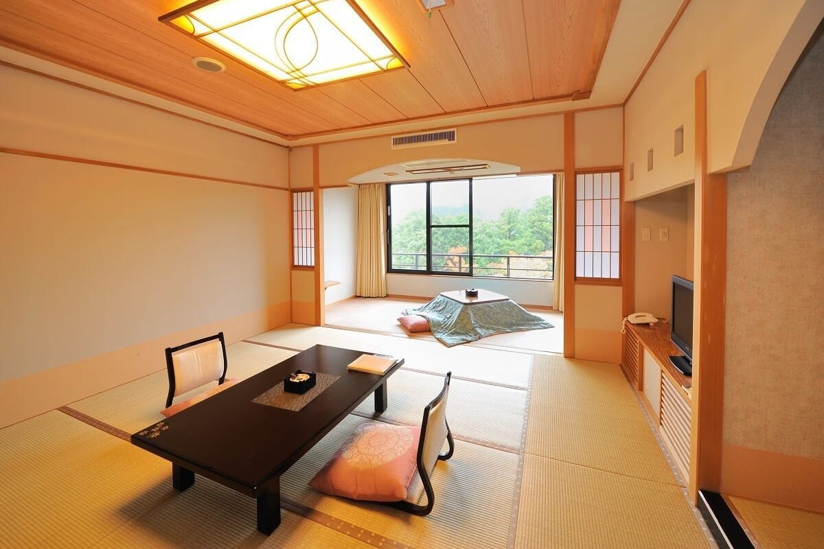 A traditional Japanese room with tatami flooring, a low table, cushions, a television, and a view of greenery outside.