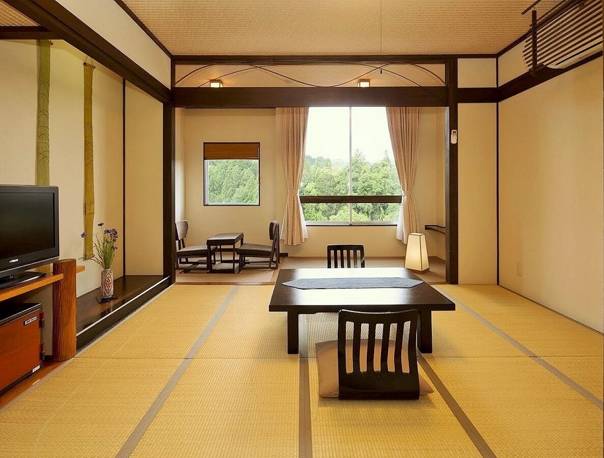 A traditional Japanese room with tatami flooring, a television, a dining table with chairs, and a view of greenery through the window.