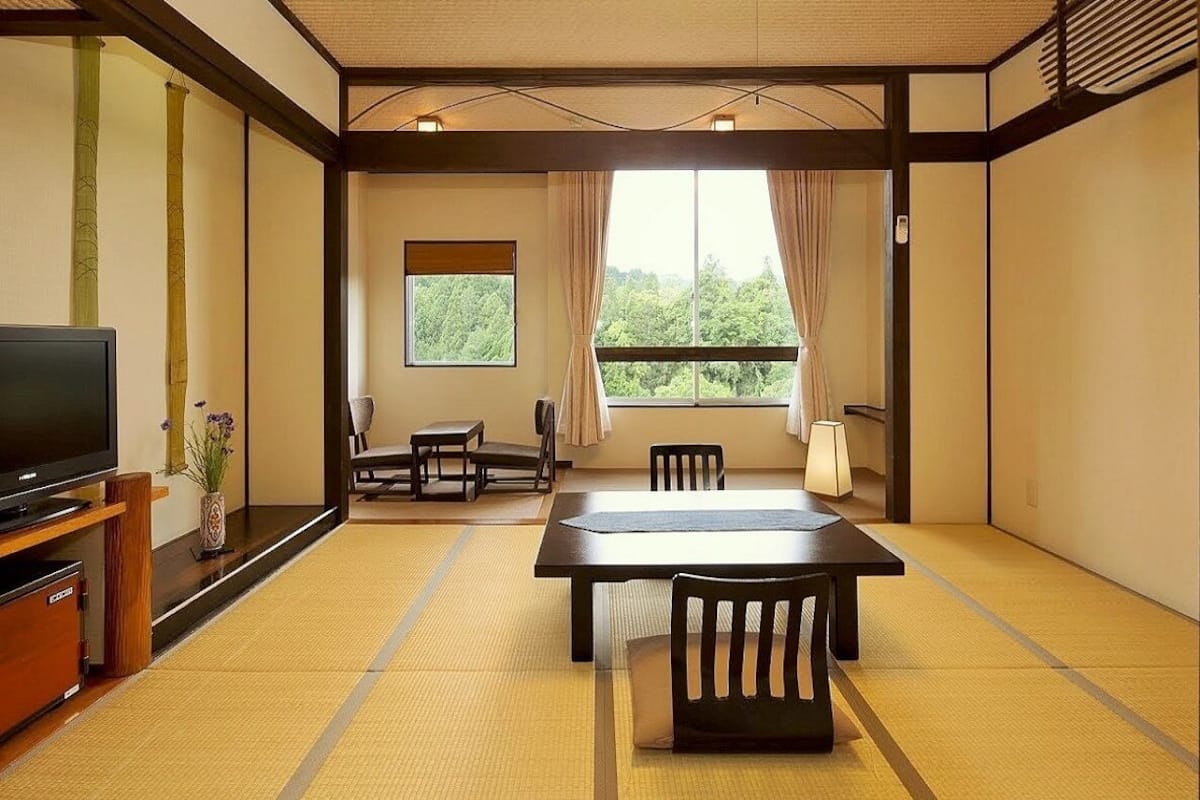 A traditional Japanese room with tatami flooring, a television, a dining table with chairs, and a view of greenery outside.