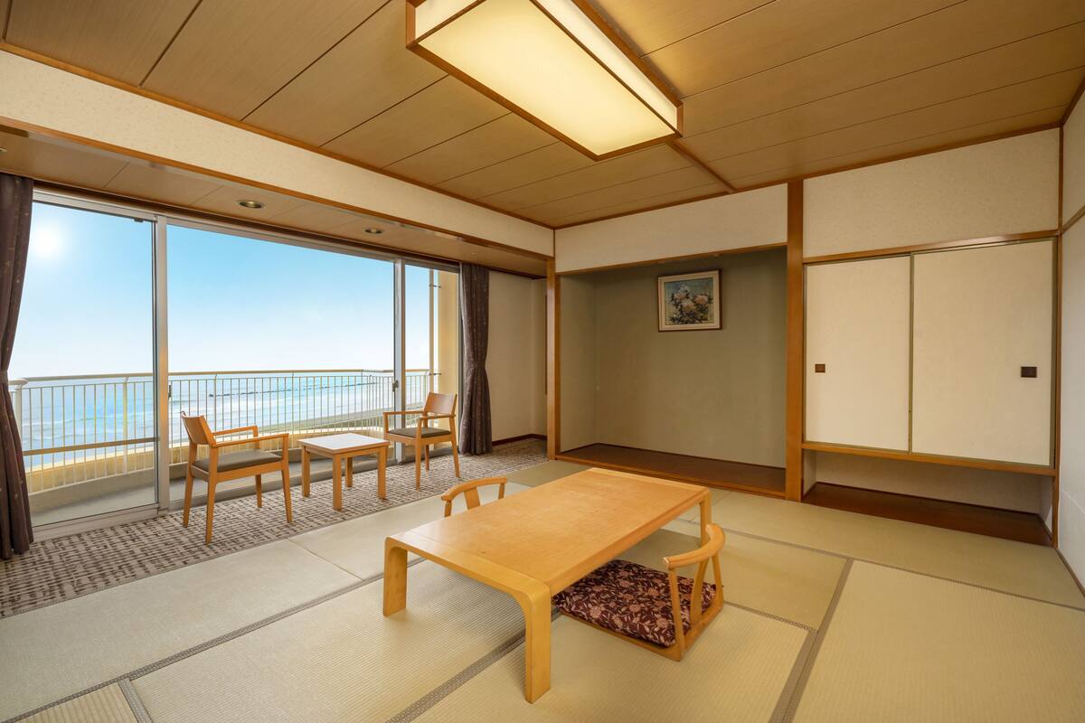 A traditional Japanese room with tatami flooring, a wooden table, chairs, a sliding door, and a view of the sea.