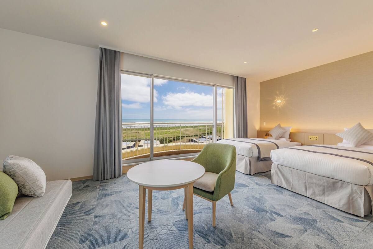 A hotel room with a large window, a round table, a green armchair, and two beds.
