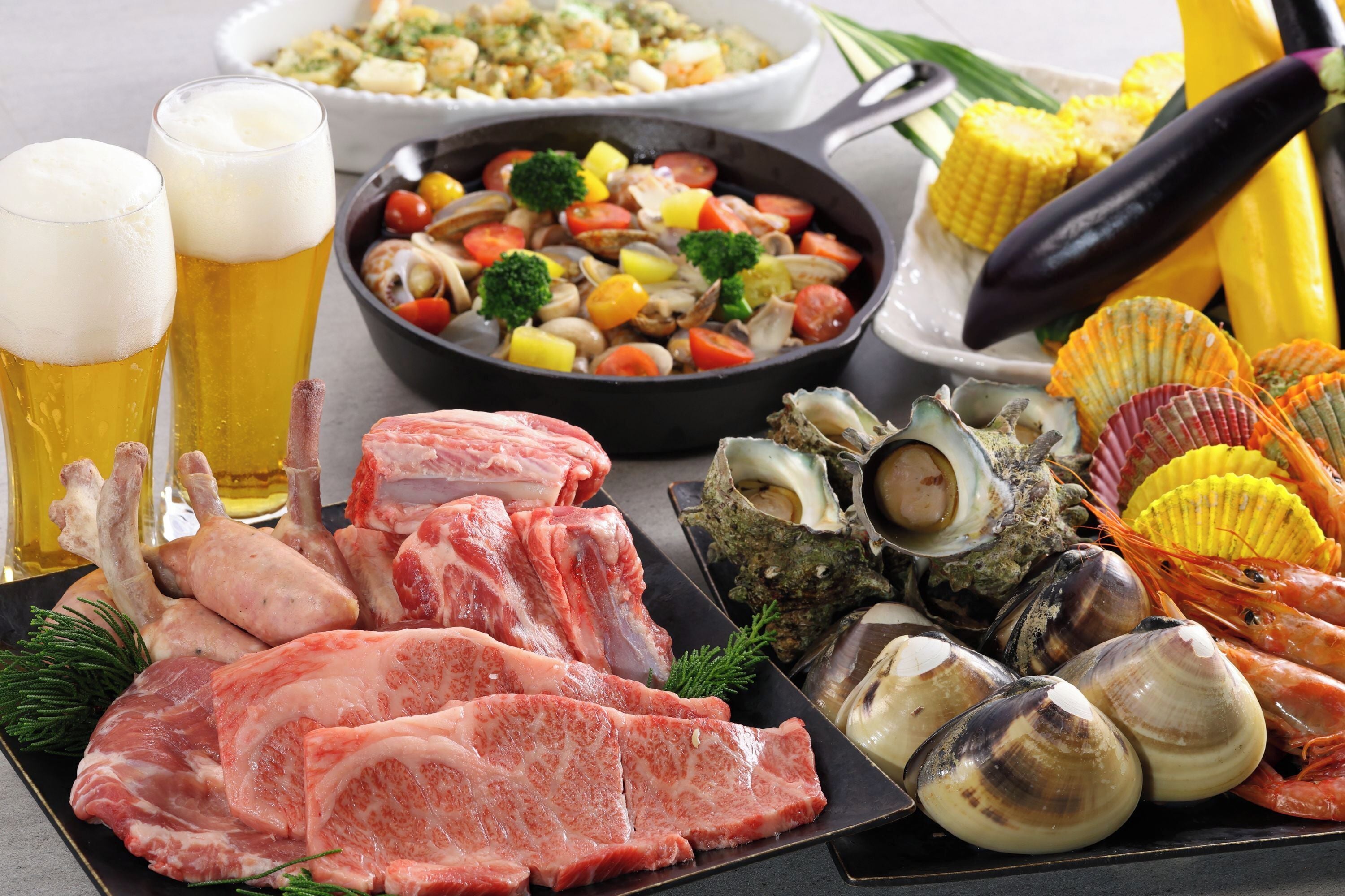 A variety of seafood and meats on a plate, a skillet with vegetables, corn on the cob, and two glasses of beer.