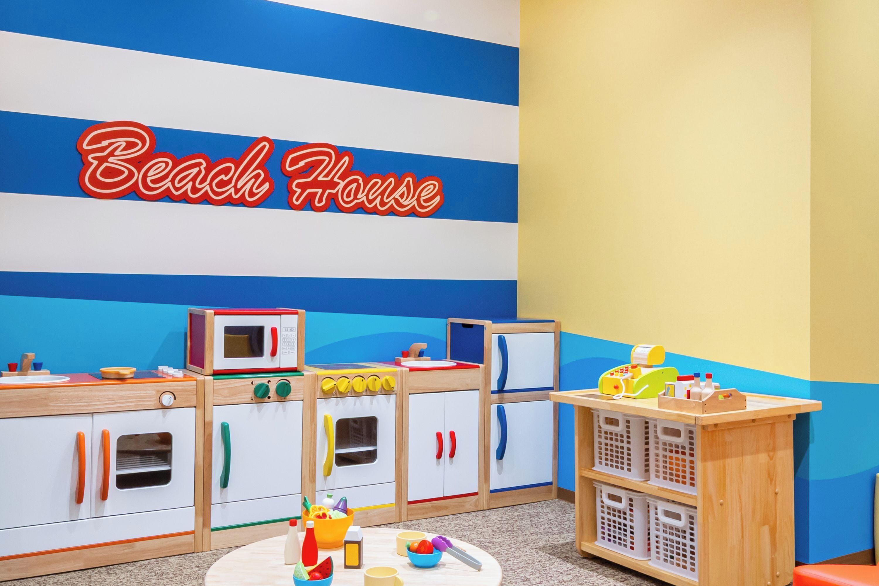 Zona infantil amb cuina de joc, taula de joc i una paret amb el rètol 'Beach House'.