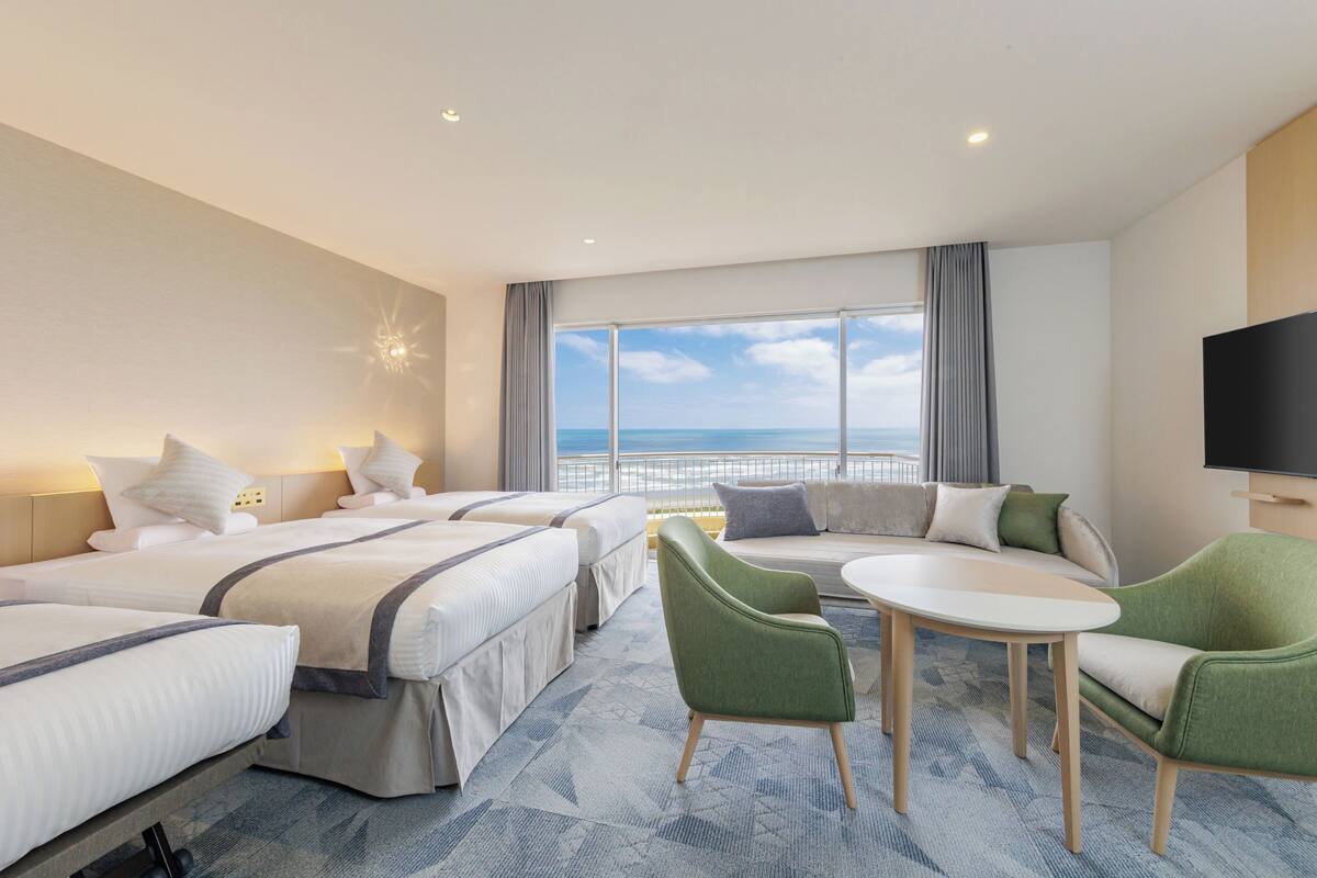 A hotel room with two beds, a sofa, a TV, and a view of the ocean.