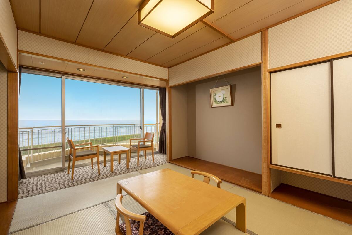 A traditional Japanese room with tatami flooring, a low wooden table, and chairs. It features a sliding glass door leading to a balcony with a view of the sea.