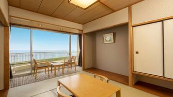 A traditional Japanese room with tatami flooring, a low wooden table, and chairs. It features a sliding glass door leading to a balcony with a view of the sea.