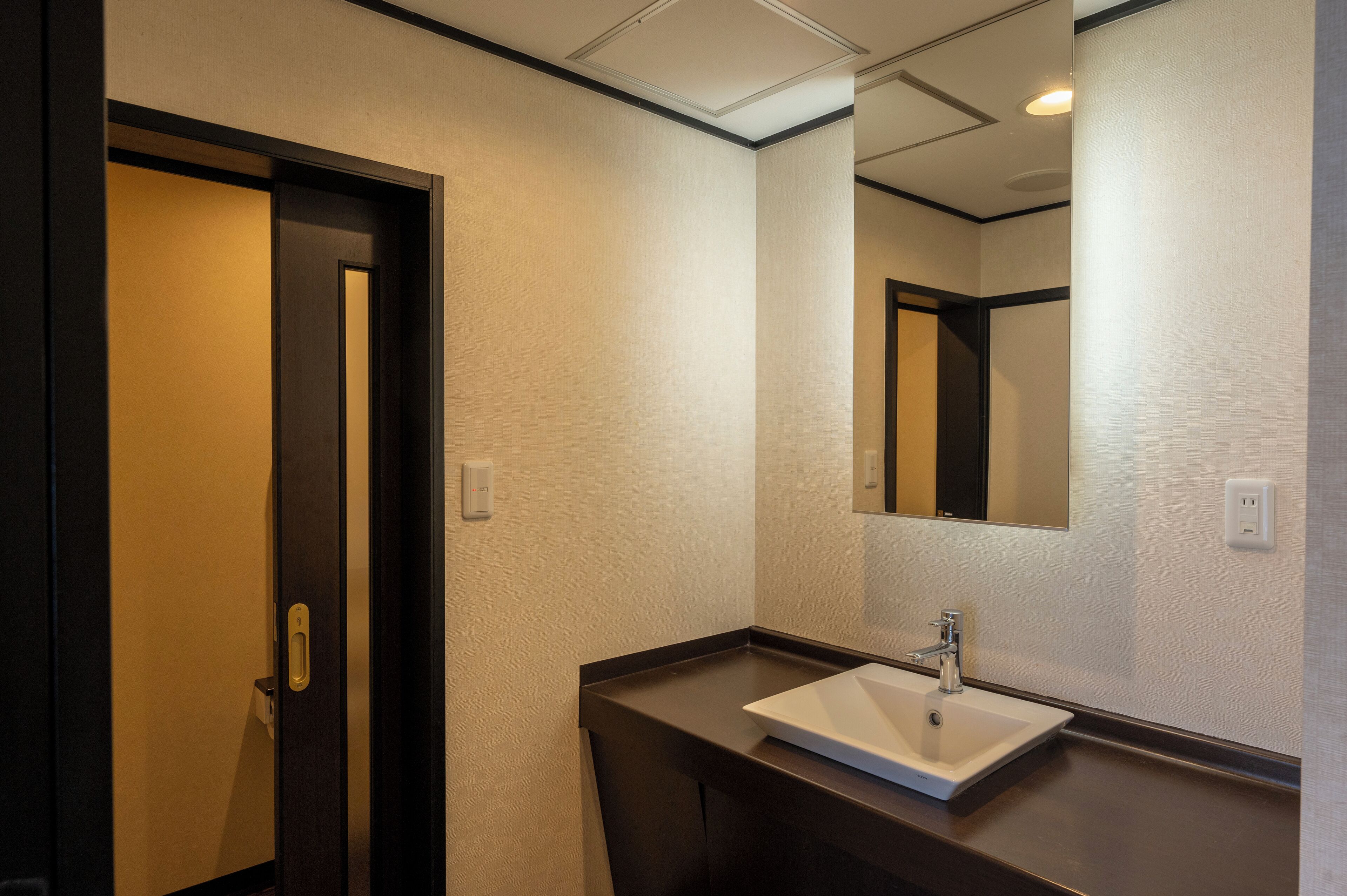 A hotel bathroom with a dark wooden door, a white sink, and a mirror on the wall.