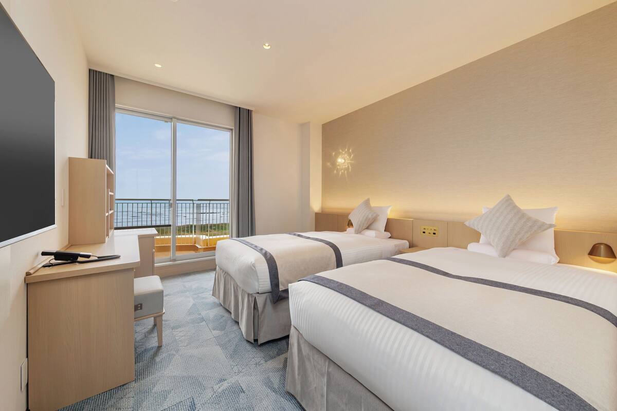 A hotel room with two beds, a desk, a chair, a large window with a view, and a balcony.