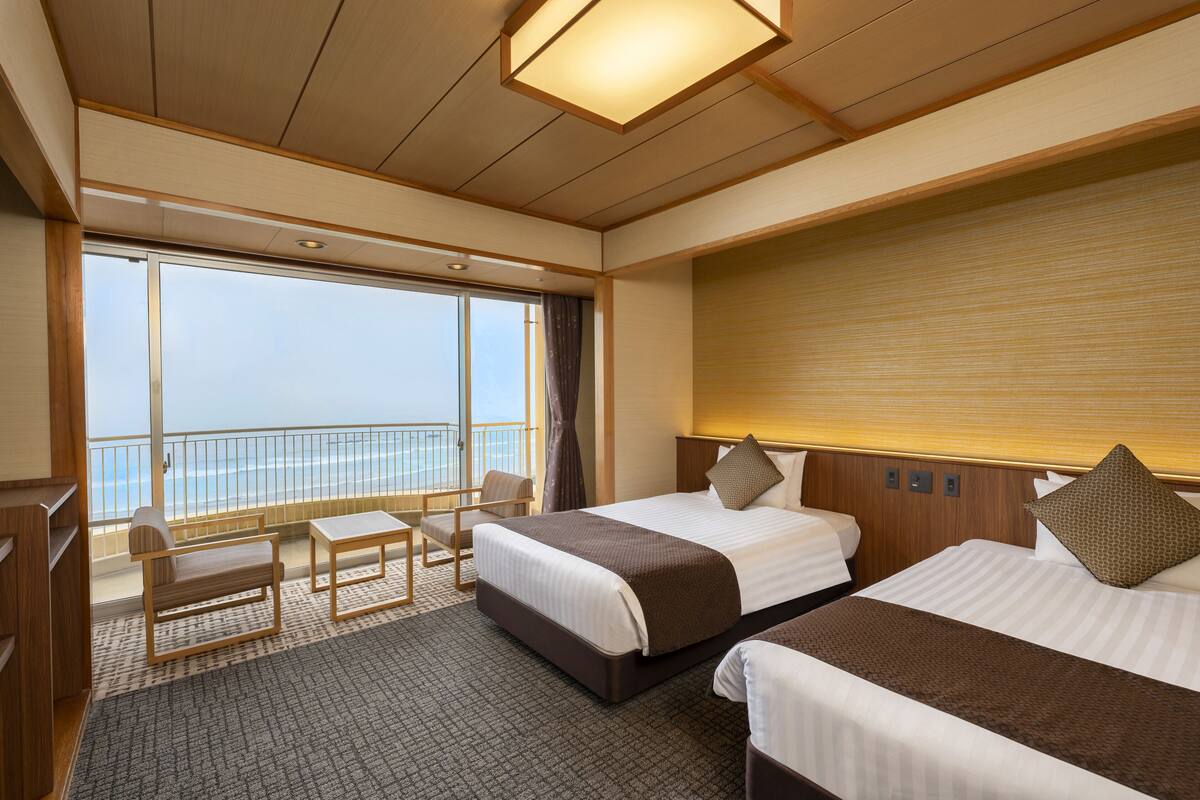 A hotel room with two beds, a desk, and a chair. There is a large window with a view of the ocean.