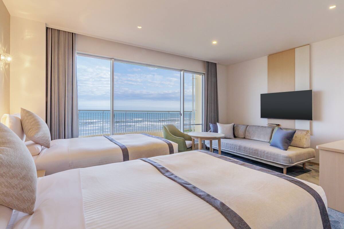 A hotel room with two beds, a sofa, a TV, and a view of the sea.